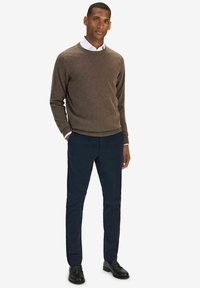 Ruskea neulepaita, tummansiniset housut ja mustat kengät. Neulepaidan kaulus on pyöreä; housuissa on sileä pinta ja hyvin istuva leikkaus.