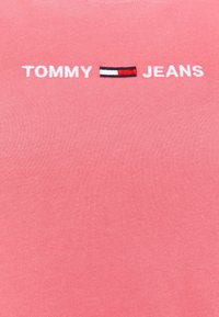 Camiseta de algodón rosa con textura acanalada. Presenta bordado "TOMMY" y "JEANS" en blanco, y un pequeño logotipo de bandera horizontal en rojo, blanco y azul.