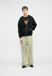 Musthave musta tõmblukuga pusakar, millel on ees tasku, koos helesiniste laiadest pükstest. Särgil on graafika kollase ja punase varjundites.