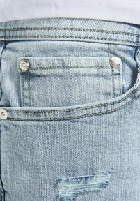 Jeans de mezclilla azul claro con un bolsillo desgastado y bordes deshilachados, herrajes metálicos y detalle de costura amarilla a lo largo de las costuras.