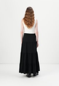 Femme aux longs cheveux bruns ondulés, portant un haut blanc sans manches et une jupe longue noire à volants, debout devant un fond blanc uni.