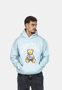 Felpa con cappuccio azzurra realizzata in tessuto morbido, caratterizzata da una grafica di un orso con il testo "Crystal Paris", grande tasca frontale e cappuccio regolabile con cordino.