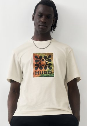 Άνδρας με σκοτεινό δέρμα και dreadlocks, φορώντας λευκό T-shirt με πολύχρωμο αφηρημένο σχέδιο και το κείμενο «HUGO», στέκεται μπροστά σε λευκό φόντο.