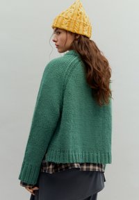 Vihreä neulottu neulepaita väljässä istuvuudessa, resorikahvoilla, käytetty ruutupaidan päällä. Keltainen neulottu pipo, jossa on teksturoitu kuvio.