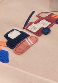 Tissu beige avec des éléments brodés colorés : un chapeau bleu, une pantoufle brune avec des rayures blanches et un véhicule rouge avec des détails en crème et marine.