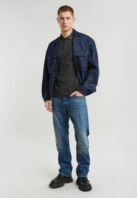 Giacca blu marina sopra una polo nera, con jeans in denim azzurro chiaro e scarpe nere chunky. La giacca presenta tasche frontali e una vestibilità rilassata.