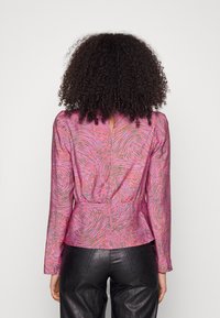 Donna con capelli ricci che indossa una blusa rosa a maniche lunghe con un motivo, pantaloni neri di pelle, girata di spalle su uno sfondo neutro.