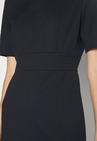 Robe noire ajustée à manches courtes, avec une taille cintrée, une texture subtile et des coutures détaillées le long des assemblages.