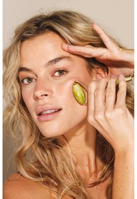 Rouleau de jade vert avec des éléments dorés, utilisé sur la joue d'une personne aux cheveux blonds ondulés et au maquillage naturel.
