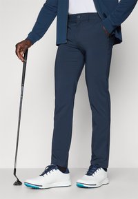 Navy golf pants in a sleek, tapered design with a smooth texture; paired with white and blue golf shoes and a black golf club.