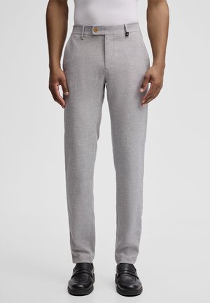 Uomo che indossa pantaloni grigi chiari con trama, mocassini in pelle nera e una camicia bianca infilata, in piedi contro uno sfondo semplice.