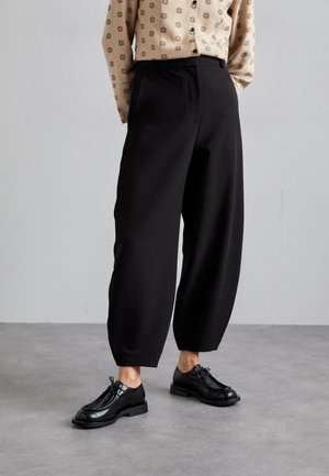 Osoba ubrana w luźne czarne spodnie, czarne sznurowane buty i beżową koszulę na guziki z ciemnymi kwiatowymi wzorami, stojąca na szarej podłodze.