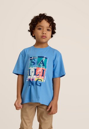 T-shirt en coton bleu clair arborant un graphique de voile coloré avec du texte. Manches courtes et encolure ronde. Porté avec un pantalon beige.