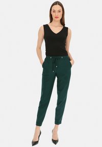 Czarny bezrękawnik z dekoltem w kształcie litery V, w połączeniu z dopasowanymi ciemnozielonymi spodniami z bocznymi kieszeniami i sznurkiem w talii. Czarne szpilki.