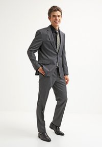 Grijze pak met een subtiele ruitpatroon, dichtgeknoopt jasje en rechte broek. Gecombineerd met een zwart overhemd en een gestreepte stropdas. Zwarte schoenen.