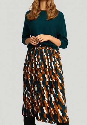 Femme portant un pull turquoise et une jupe plissée taille haute avec un motif géométrique en marron, noir, blanc et turquoise.