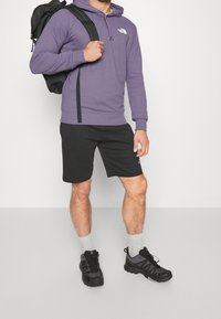 Lila huvtröja med känguruficka, svarta shorts och svarta träningsskor. Har en logotyp på huvtröjan och ryggsäckstroppar.