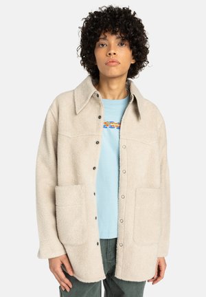 Beige fleece shirtjack met een gekraagde hals, zwarte knopen, twee voorzakken en een zachte textuur. Draagt over een lichtblauw shirt.