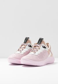 Paire de baskets de sport rose pâle et beige avec détails perforés, superpositions blanches ondulées, lacets à motifs et languettes de tirage noires.