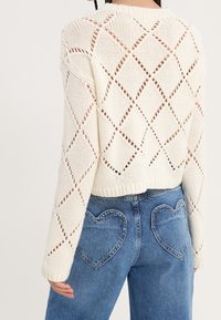 Crèmekleurig gebreid sweater met diamantpatronen en geribde onderrand, gecombineerd met blauwe jeans met hartvormige achterzakken.