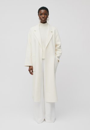 Femme aux cheveux courts portant un long manteau blanc, un pull à col roulé blanc et un pantalon blanc large, debout devant un fond clair uni.