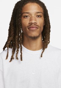 Moški z medijem rjavimi dreadlocksi in svetlo brado, oblečen v belo jopico Nike z majhnim logotipom na prsih.