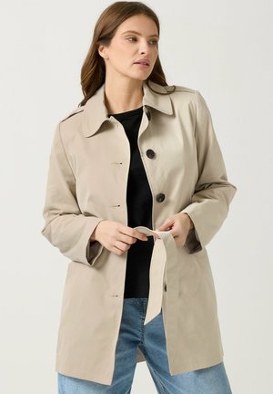 Femme attachant une ceinture sur un trench-coat beige porté sur un haut noir et un jean bleu, sur fond clair uni.