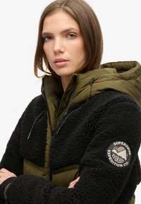Zwarte fleecejack met een olijfgroene gewatteerde binnenvoering en capuchon. Voorzien van een logopatch op de linkermouw en een rits aan de voorkant met koordjes.
