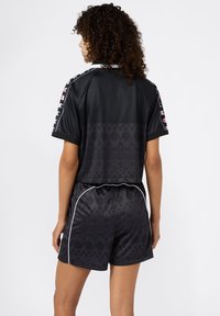Donna con capelli ricci che indossa pantaloni sportivi neri a fantasia e una maglietta a maniche corte coordinata con strisce e logo, rivolta lontano dalla telecamera.