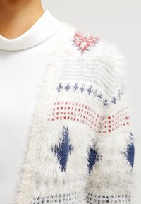 Bílý chlupatý kardigan s červenými a modrými geometrickými vzory, s měkkou texturou a otevřeným předním designem.