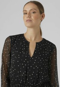 Robe noire en tissu léger avec de longues manches transparentes, décolleté en V et motif à pois de couleurs claires, taille froncée.