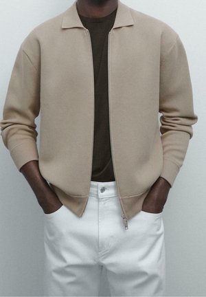 Osoba ubrana w beżową kurtkę na zamek błyskawiczny nałożoną na ciemnobrązową koszulę, z rękami w kieszeniach białych spodni, na jasnym, jednolitym tle.