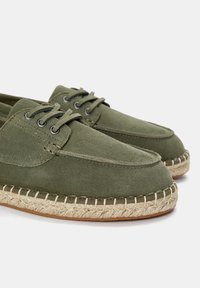 Maslinasto zelene espadrile s gornjom od antilopa, okruglim vrhom i šivenim jute potplatom. Ima šest rupica s tamnim vezicama.