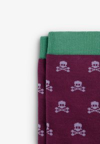 Calcetines morados con un puño verde, que presentan un patrón repetido de calaveras y huesos cruzados en gris claro. Textura de punto en toda su superficie.