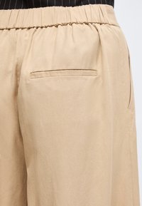 Μπεζ παντελόνι με ελαστική μέση και μία μόνο πίσω τσέπη με ψαλιδωτή ραφή, που φοριέται με μαύρο τοπ με λεπτές ρίγες.