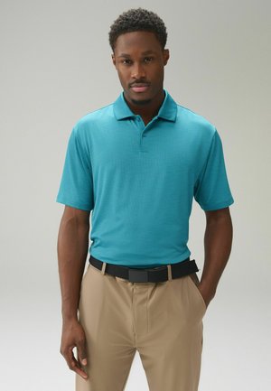 Mand iført en kortærmet polo i turkis, stoppet ned i beige bukser med sort bælte, stående mod en ensartet lys baggrund med hænderne i lommerne.