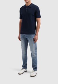 Marineblauwe polo met korte mouwen en een textuur gebreide patroon, drie knopen bij de kraag, gecombineerd met lichtblauwe jeans en witte sneakers.