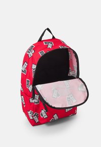 Mochila roja con patrón de logotipos "NIKE" en blanco y negro. Presenta una abertura con cremallera y un forro interior negro con un acento rosa.