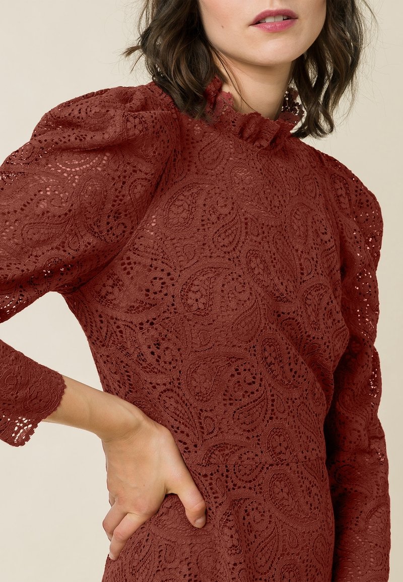 Robe en dentelle de couleur rouille avec un motif paisley, un col haut à volants et des manches bouffantes aux bords festonnés.