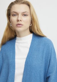 Helle blaue Strickjacke, offene Vorderseite, aus weichem Material. Getragen über einem weißen T-Shirt, das einen lässigen Schnitt und eine glatte Textur aufweist.