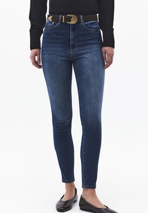 Mujer con vaqueros ajustados azul oscuro, cinturón negro con hebilla dorada, camiseta de manga larga negra y zapatos planos negros, de pie sobre un fondo blanco.