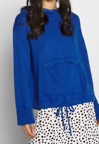 Blå stickad hoodie med en framficka, dragsko i nederkant och ribbade ärmslut, tillsammans med en prickig kjol.