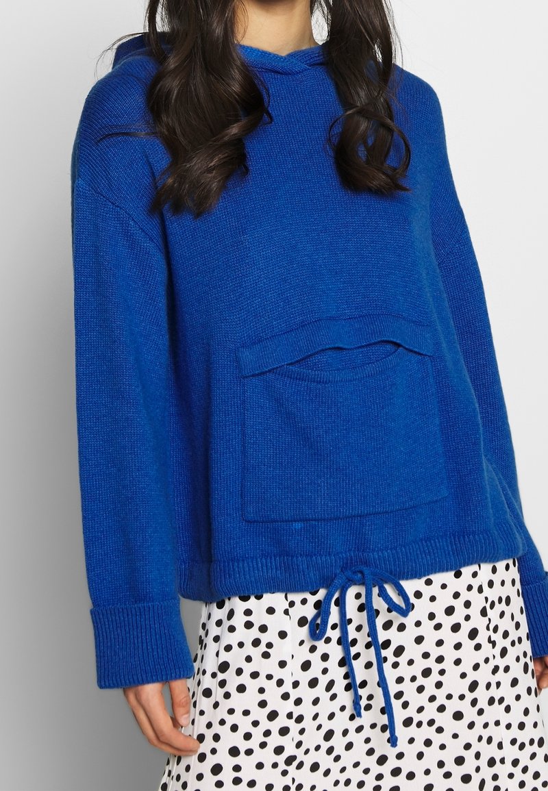 Blå stickad hoodie med en framficka, dragsko i nederkant och ribbade ärmslut, tillsammans med en prickig kjol.