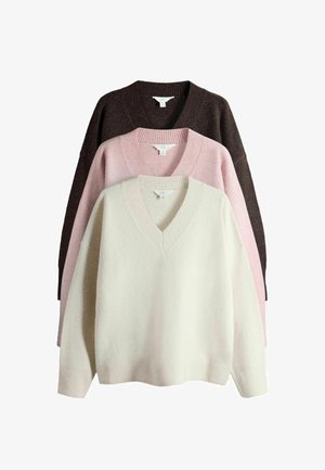 Frau mit langen dunklen Haaren, die einen locker sitzenden schwarzen V-Ausschnitt-Pullover, weite schwarze Spitzenbesetzte Hosen und schwarze offene Sandalen trägt, steht vor einer weißen Wand.