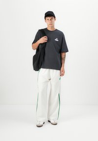Sort kasket, grå oversized t-shirt med logo, hvide bukser med vidde og grønne striber, samt hvide sneakers med grå detaljer.