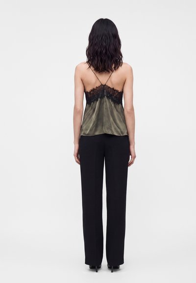 Femme debout, tournée vers l’arrière, portant un caraco en satin vert olive avec de la dentelle noire et un pantalon large noir sur fond uni.