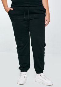 Persoon draagt zwarte joggingbroek met elastische boorden en witte chunky sneakers, staand met handen in de zakken tegen een effen achtergrond.