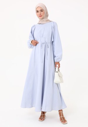 Nainen, joka pukeutunut vaaleansiniseen pitkähihainen mekkoon ja beigeen hijabiin, pitää valkoista käsilaukkua, seisoo valkoisissa remmi-koroissa yksinkertaisella taustalla.