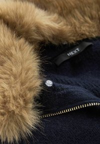 Svart ullrock med gulddragkedja och beige konstpälskrage, visar en klar plastknapp och svart klädbetäckning med texten "NEXT".