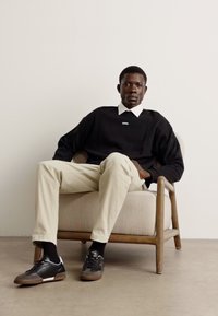 Maglione nero sopra una camicia bianca con colletto, pantaloni beige e sneakers nere seduto su una sedia beige chiaro con gambe in legno contro uno sfondo neutro.